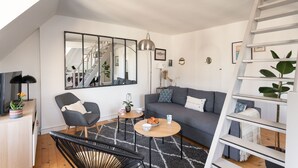 Een moderne woonkamer met een grijze bank, een zwarte stoel, een houten salontafel en een zwarte rieten stoel. Een trap met een witte leuning en een plant aan de rechterkant.