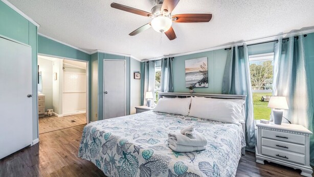 A bedroom with a bed, a ceiling fan, a nightstand, a lamp, and a view of the outdoors. - Bayside Haven by Seaside Vacations & Sales (Chincoteague Island)
