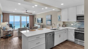 A modern kitchen with white cabinetry, a dishwasher, and a stainless steel oven. There is a large island with a sink and a dining area with chairs.