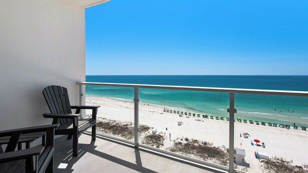 A balcony with two Adirondack chairs, a view of a beach with umbrellas, and a clear blue sky.