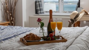 A breakfast tray with croissants, strawberries, a bottle of champagne, and glasses of orange juice on a bed with a view of the sea.