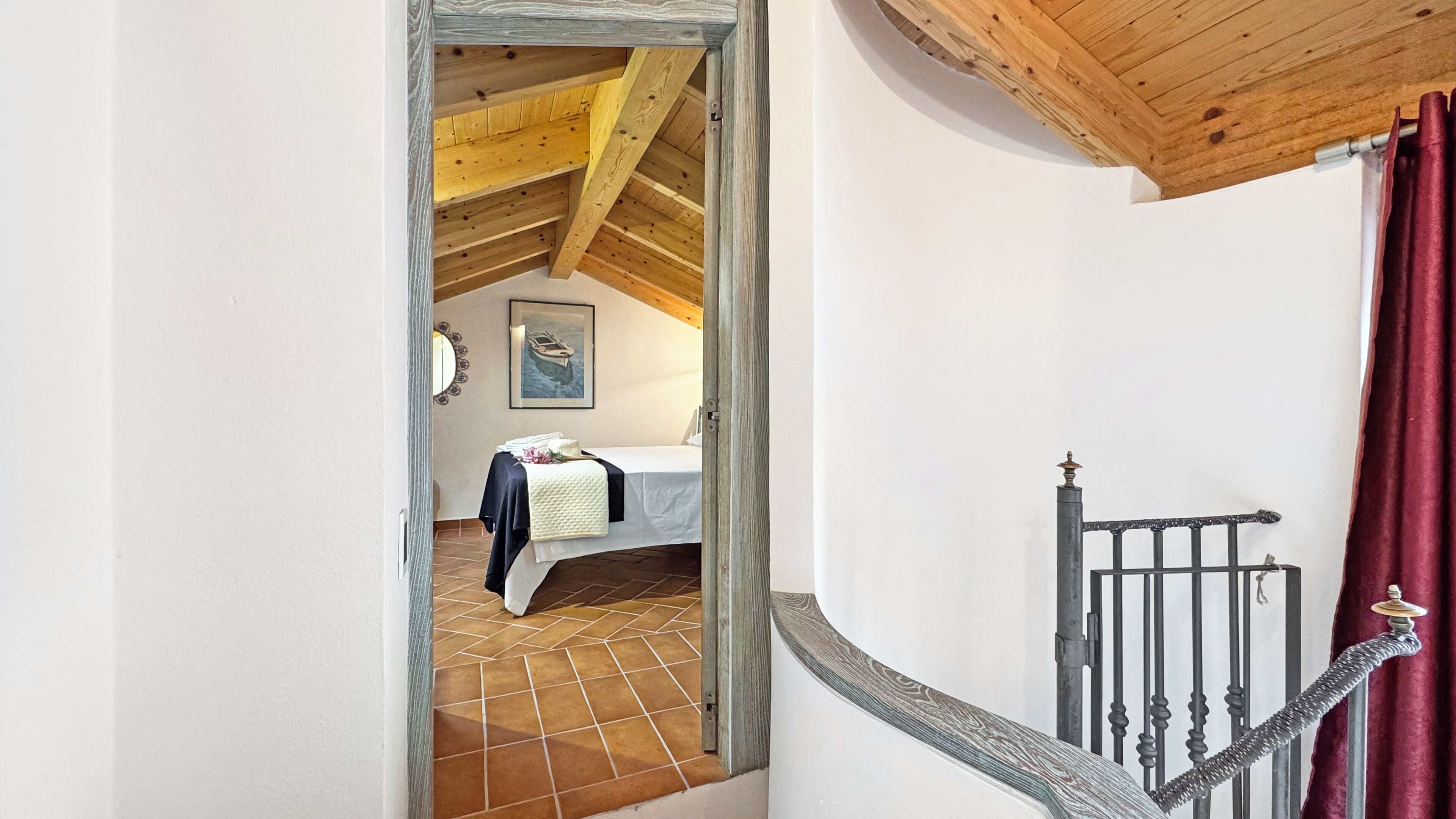 Une chambre avec un plafond en bois, un lit recouvert d’une courtepointe blanche, une petite table sur laquelle se trouve un vase, et un tableau accroché au mur.