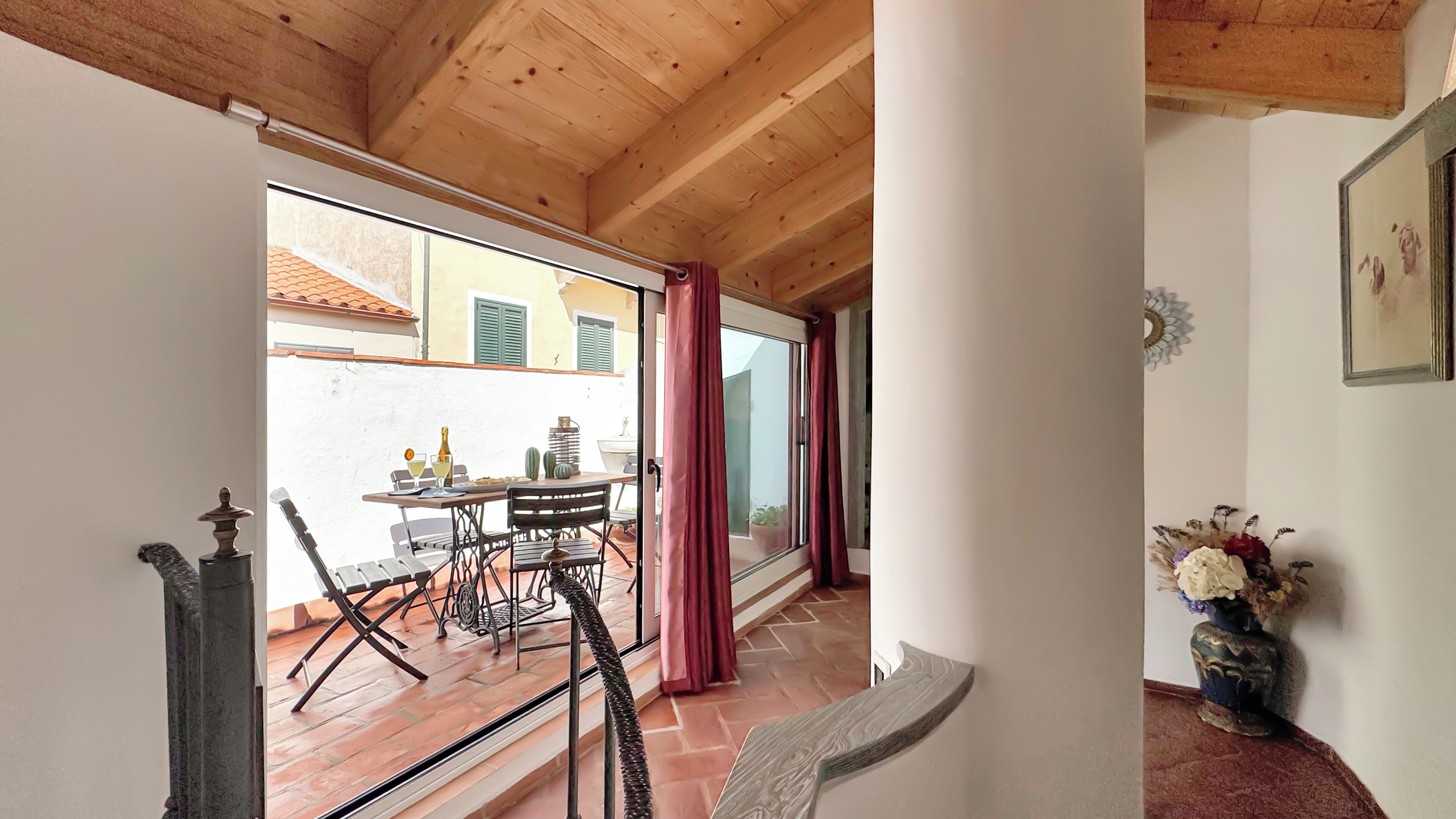 Una habitación con techo de madera, un ventanal corredero que da a un espacio exterior con una mesa y sillas, y un jarrón con flores a la derecha.