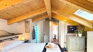 Ein gemütliches Schlafzimmer im Dachgeschoss mit geneigter Holzbalkendecke, einem Bett mit weißen Leinentüchern, einem Schreibtisch mit Lampe und einem Fenster mit Blick.