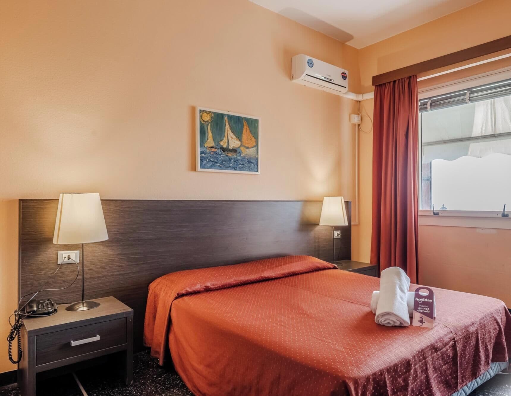 A hotel room with a bed, two bedside lamps, a painting, an air conditioning unit, a window with curtains, a telephone, and a folded towel on the bed.