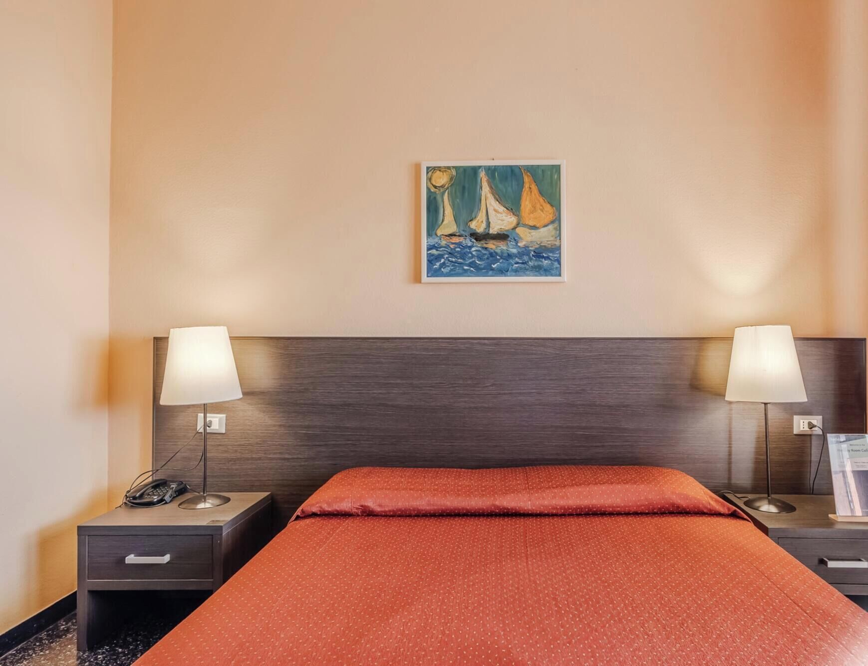 Een hotelkamer met een bed, twee nachtkastjes, een schilderij van zeilboten, en een lamp op elk nachtkastje.