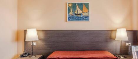 A hotel room with a bed, two bedside tables, a nightstand, a painting of sailboats, and a lamp on each table.