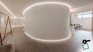A modern, minimalist room with a curved white wall and a circular light feature. There are chairs arranged in a row, and a window provides natural light.