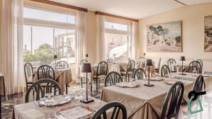 A dining area with tables set for meals, large windows with curtains, and a painting on the wall.