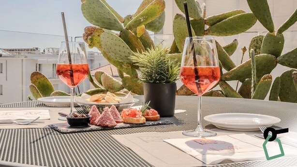 A table set with two glasses of rosé wine, a small potted plant, and a variety of appetizers including salami and cheese on bread, on a rooftop terrace with cacti in the background.