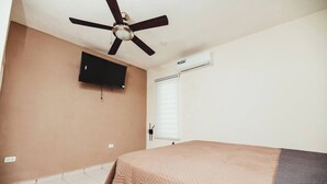 Un dormitorio con cama, ventilador de techo, televisión, aire acondicionado y ventana con persianas.