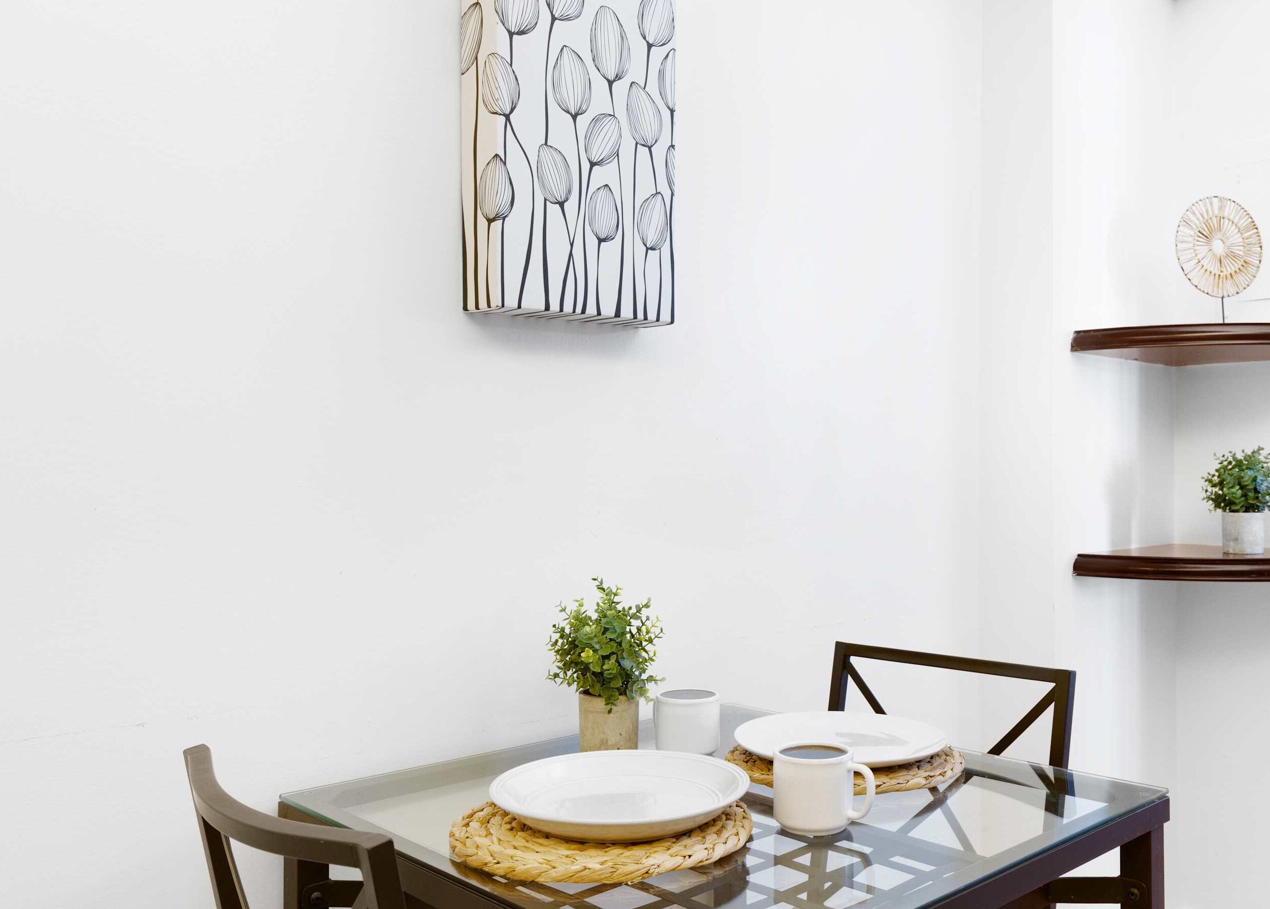 Comedor con una mesa de vidrio, individuales tejidos, platos blancos y una pequeña planta en maceta.