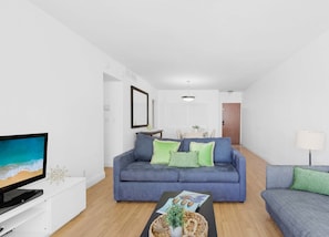 A modern living room with a blue sofa, a television, and a dining area in the background. - Beautiful APT w/ Ocean View and Beach Access (Hollywood)