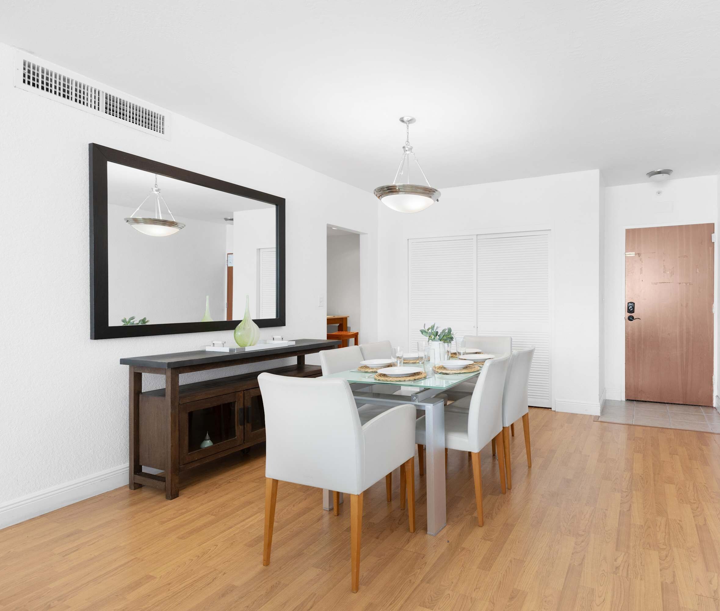 A dining area with a glass table, white chairs, a dark wooden console, and a large mirror on the wall.