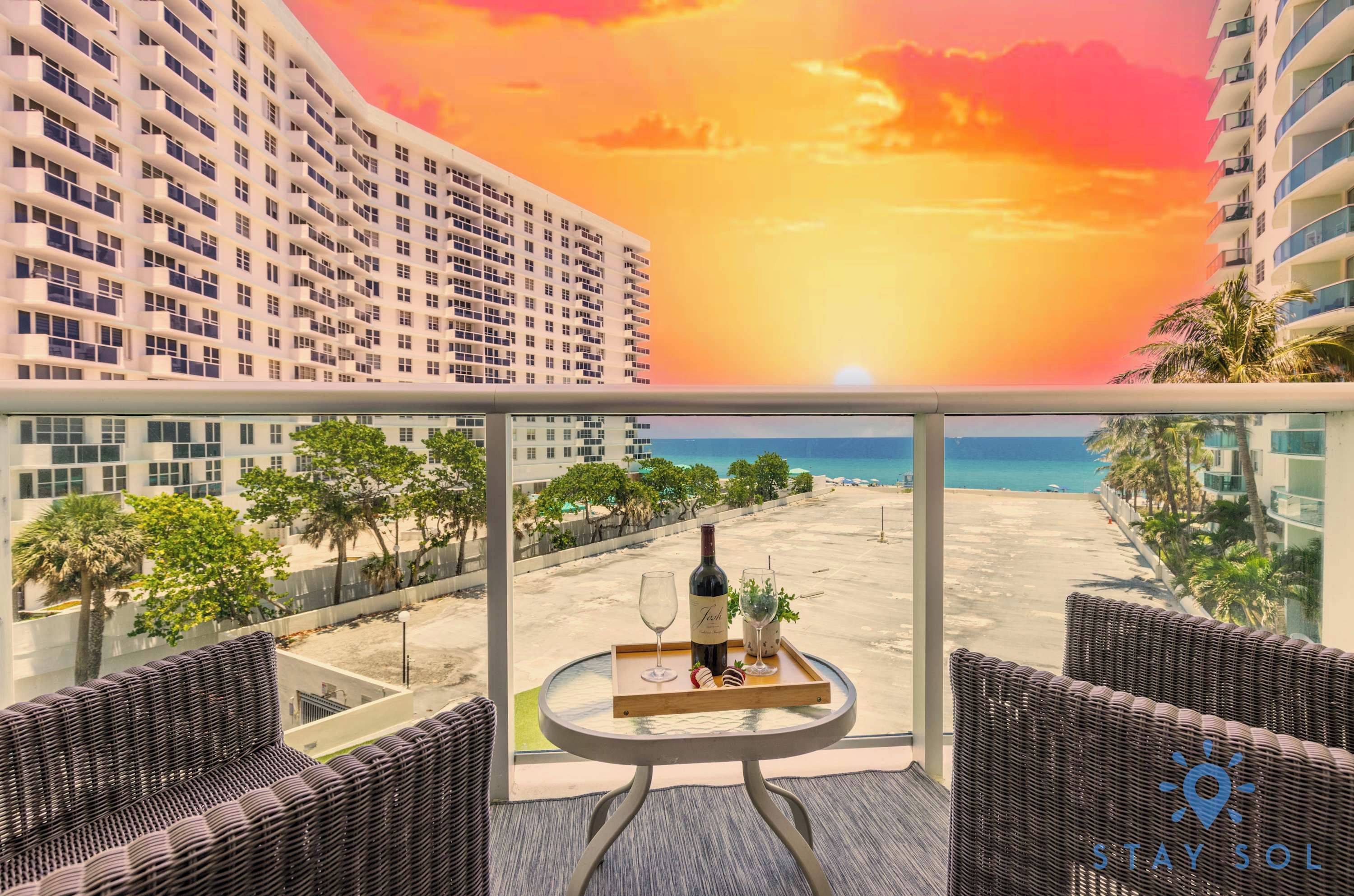 A balcony with a view of a beach and sunset, a bottle of wine, and a tray with glasses.