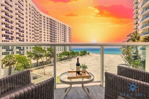 Un balcon avec vue sur la plage et le coucher de soleil, une bouteille de vin et un plateau avec des verres.