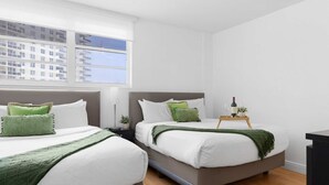 Een hotelkamer met twee bedden, een raam met uitzicht op gebouwen, een nachtkastje met een lamp, en een dienblad met een fles en glazen.