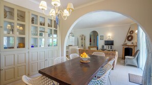 Un espace repas avec une longue table, des chaises blanches, un lustre et, à travers une arche, une vue sur un salon doté d’une cheminée.