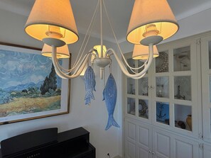 Un lustre à plusieurs bras et abat-jours, un tableau encadré au mur et un piano au premier plan.