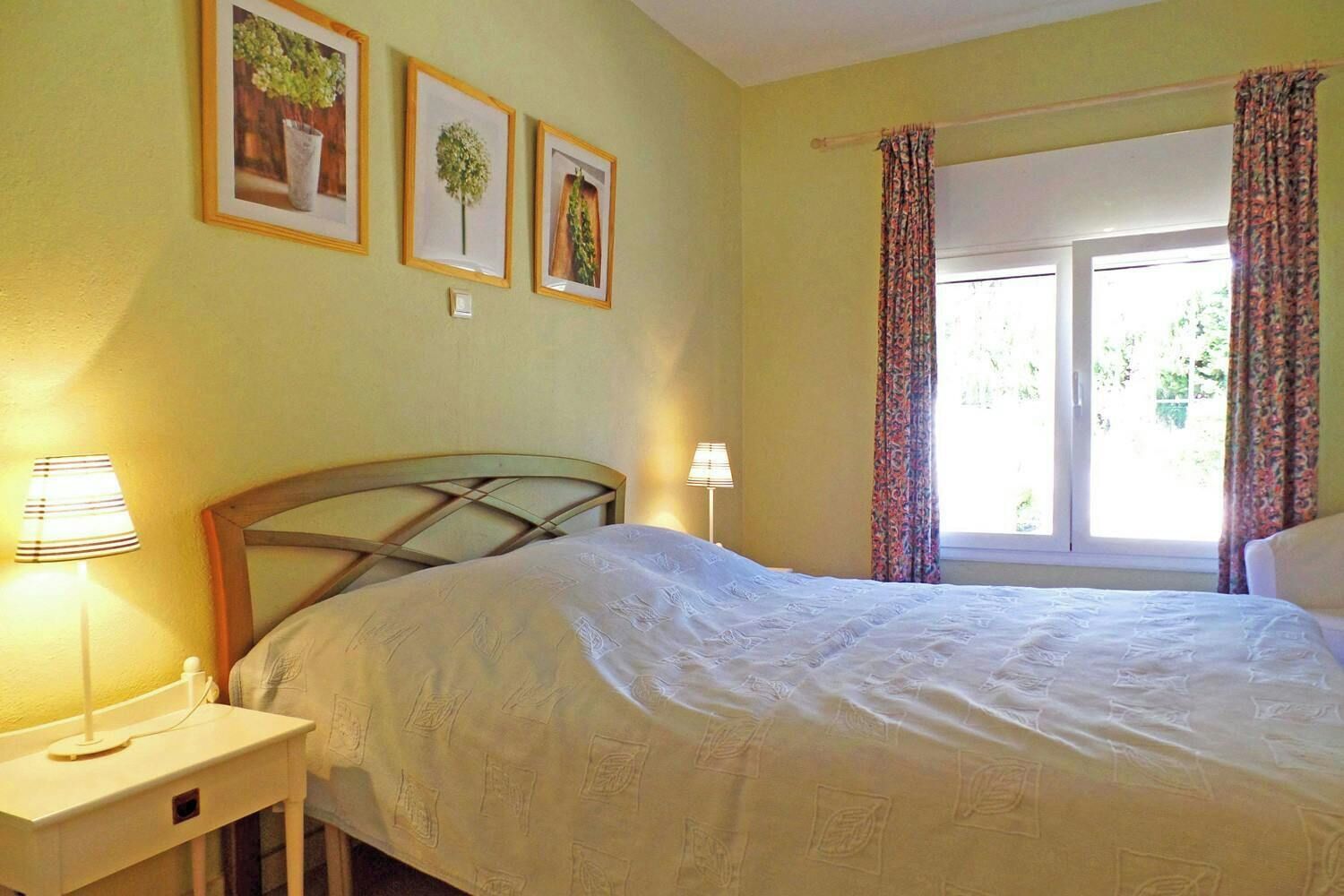 Une chambre à coucher avec un lit, deux lampes de chevet, une fenêtre avec des rideaux et trois tableaux encadrés accrochés au mur.