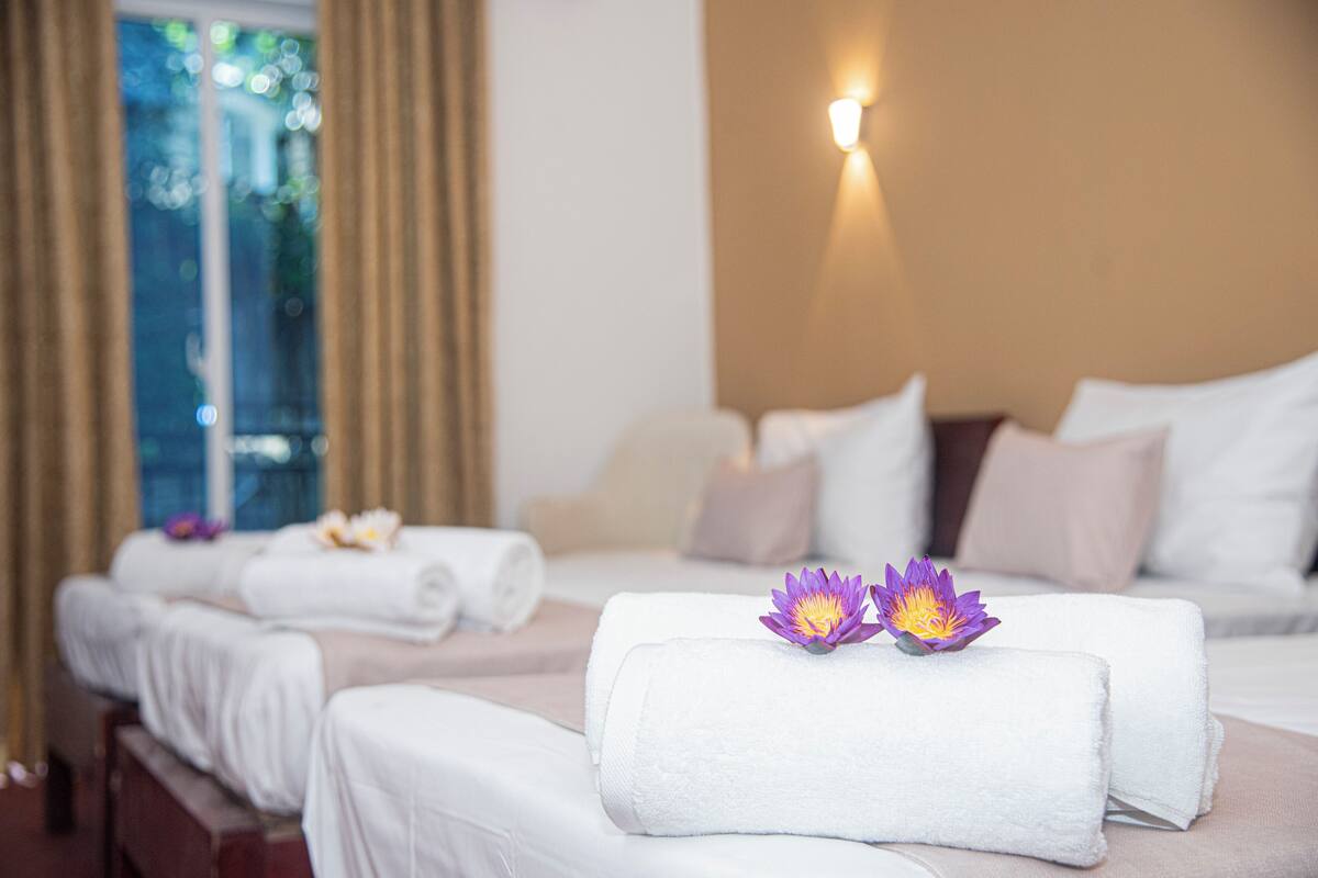 Una camera d'albergo con due letti, ognuno con un asciugamano bianco arrotolato e un fiore di loto viola.