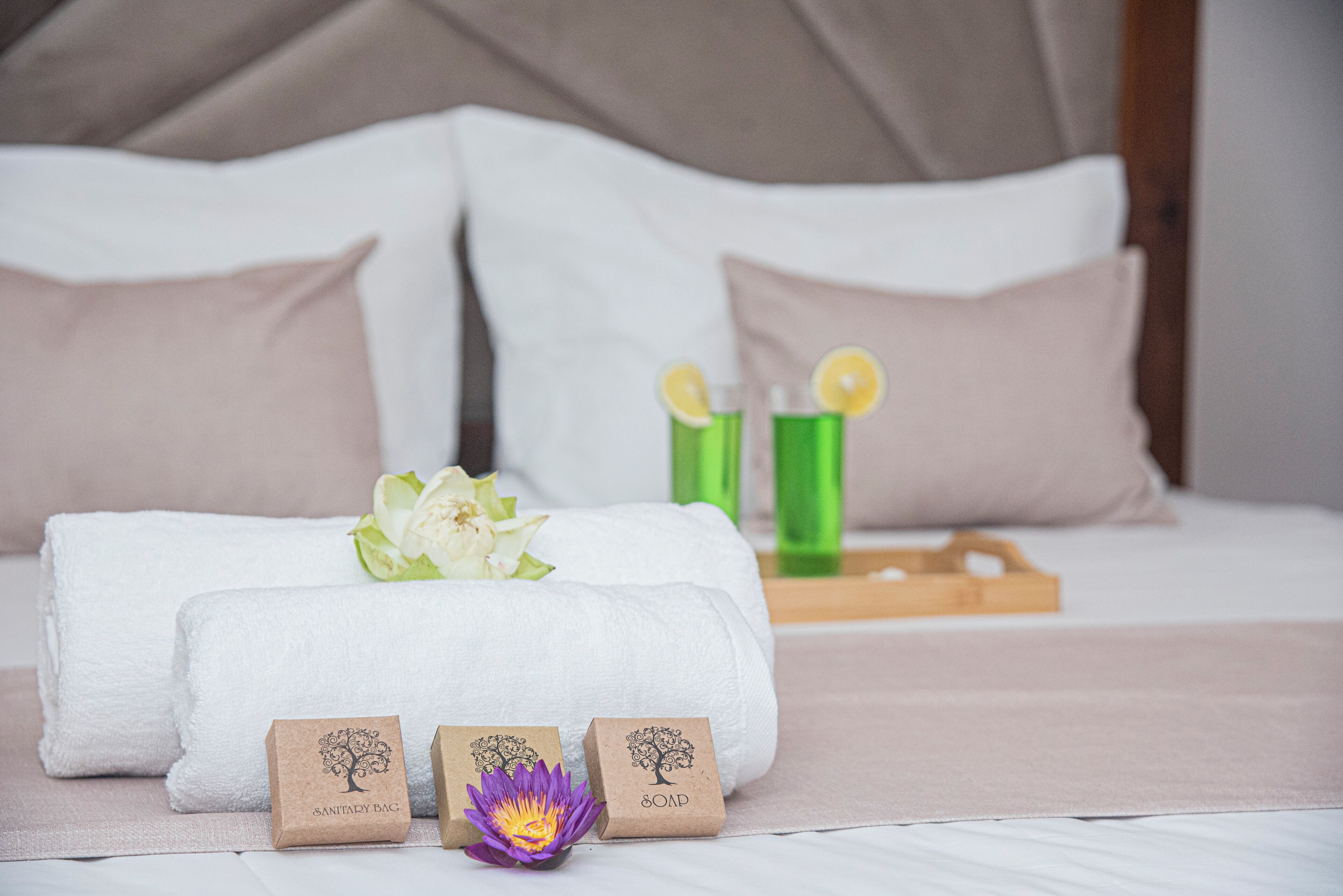 Una cama bien hecha con ropa de cama blanca y beige, una bandeja de madera con vasos verdes y dos cajas pequeñas marrones con las etiquetas 'SANITARY BAG' y 'SOAP'.
