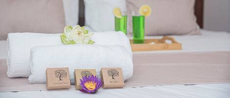 Una cama bien hecha con ropa de cama blanca y beige, una bandeja de madera con vasos verdes y dos cajas pequeñas marrones con las etiquetas 'SANITARY BAG' y 'SOAP'.