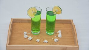 Zwei Gläser grüner Flüssigkeit auf einer Holzschale mit weißen Blumen.