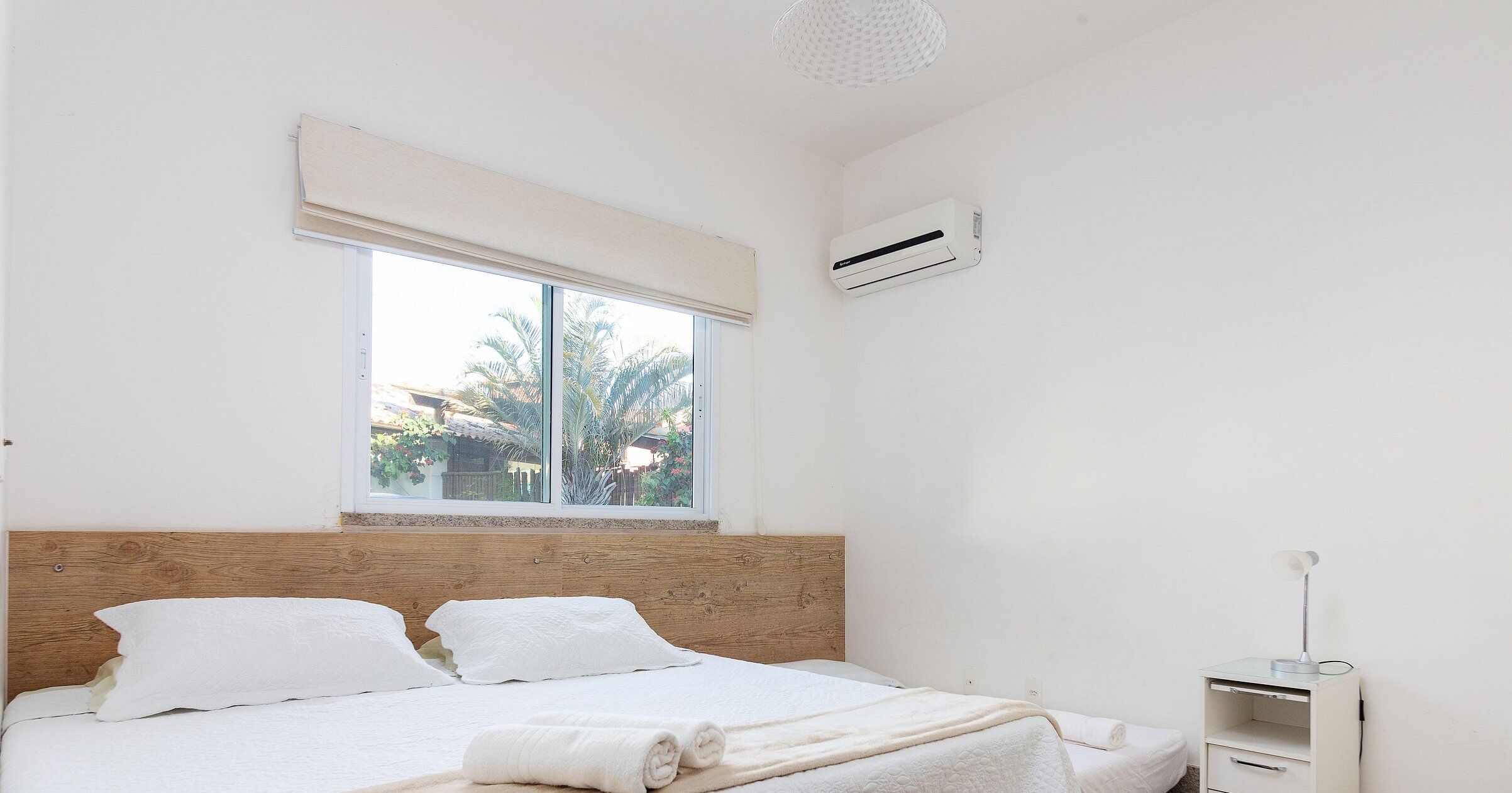 Une chambre avec une tête de lit en bois, une grande fenêtre donnant sur des palmiers, un climatiseur blanc et une table de chevet avec une lampe.