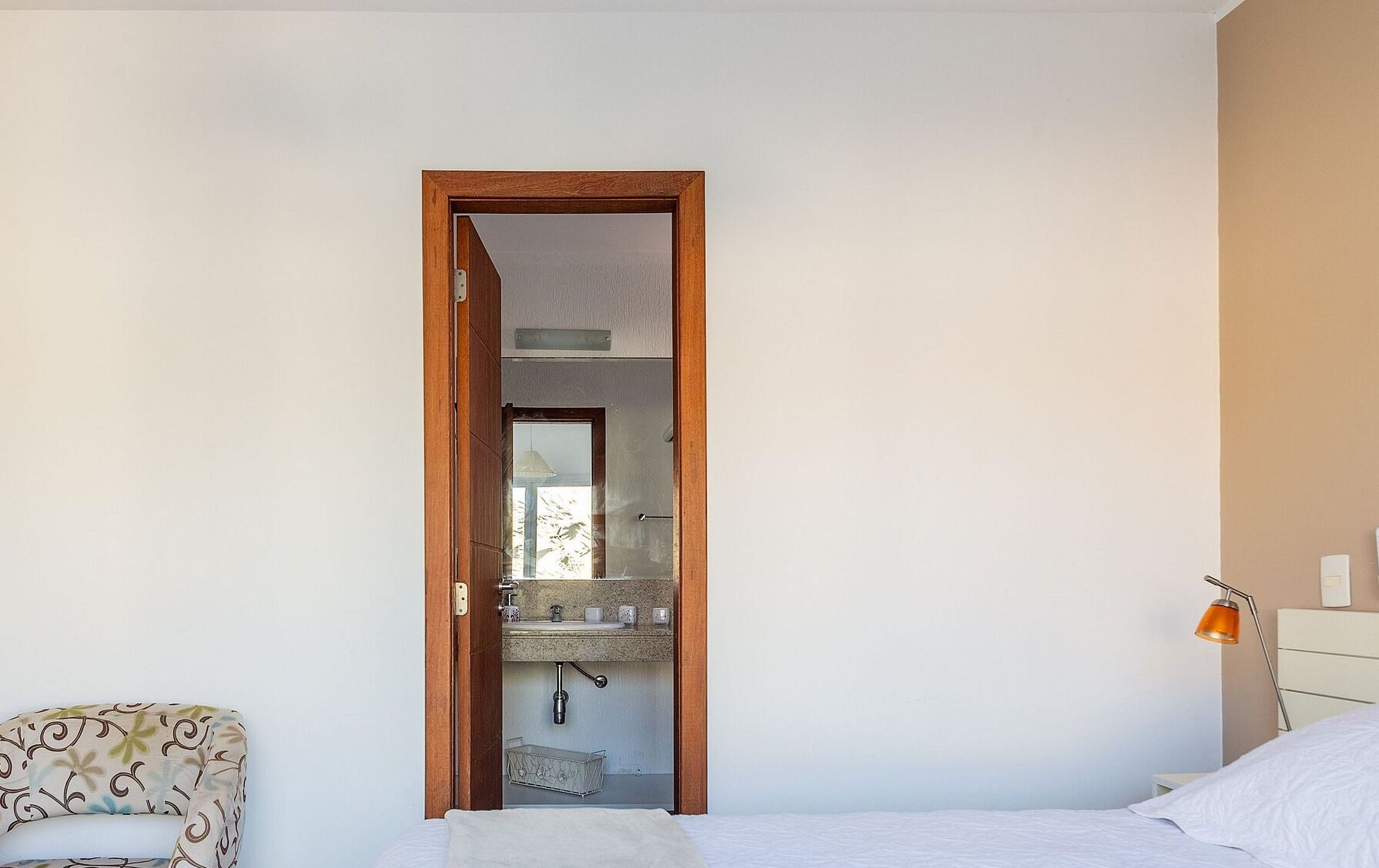 Une chambre à coucher comprenant un lit, une chaise, une porte en bois donnant sur une salle de bain, et une table de chevet avec une lampe.