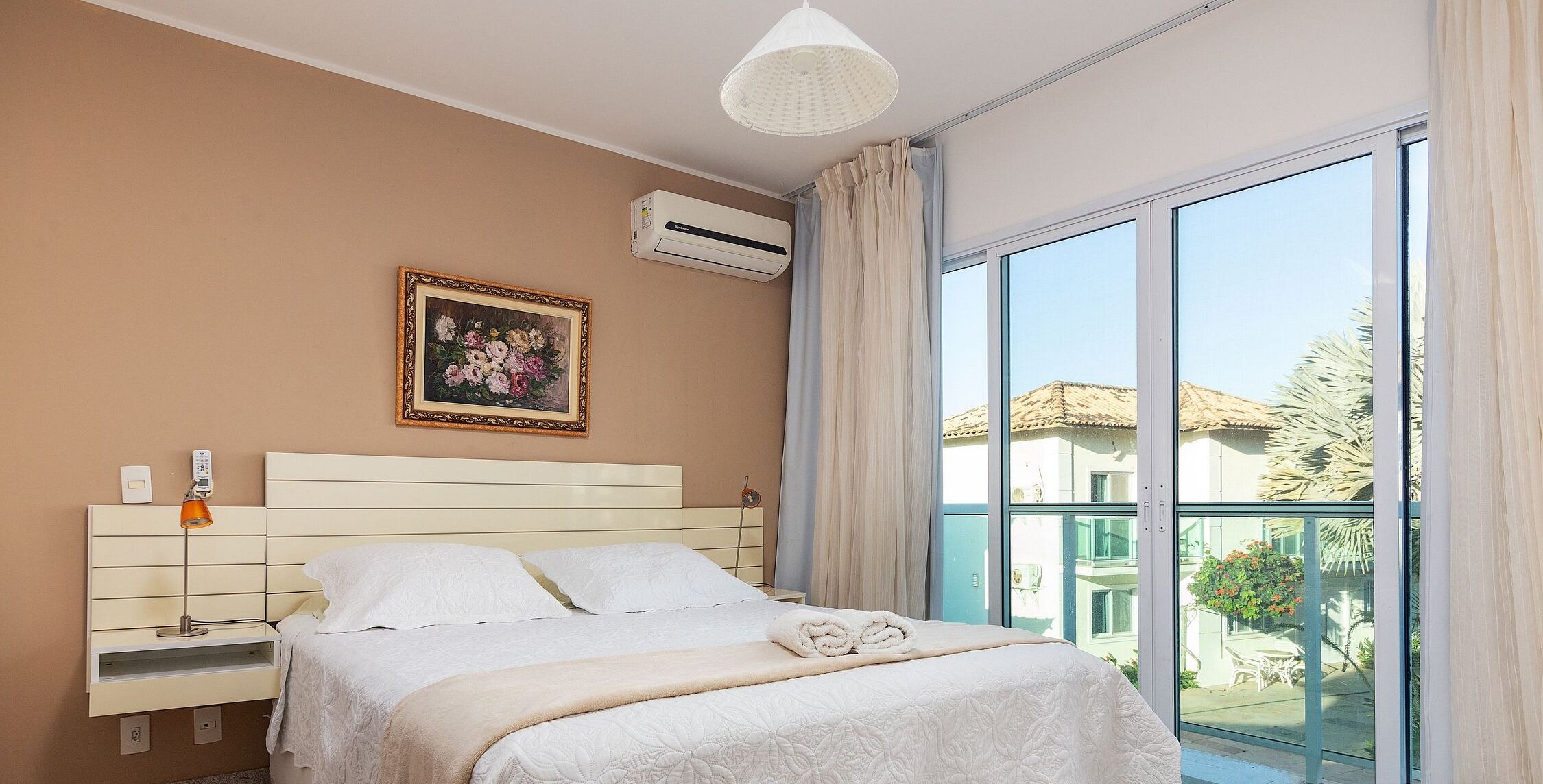 A bedroom with a bed, a bedside table, a lamp, an air conditioner, a window with curtains, and a painting on the wall.