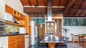 A modern kitchen with wooden cabinetry, a dining area with a wooden table and chairs, and a view of the outdoors through large windows. - Bz35 Casa com piscina aquecida em Geribá (Buzios)