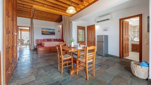 A spacious living area with a wooden dining table, chairs, and a sofa. A kitchen area is visible with a refrigerator and a stone wall. - Arte Pefki Beachfront Suites (Rhodes)