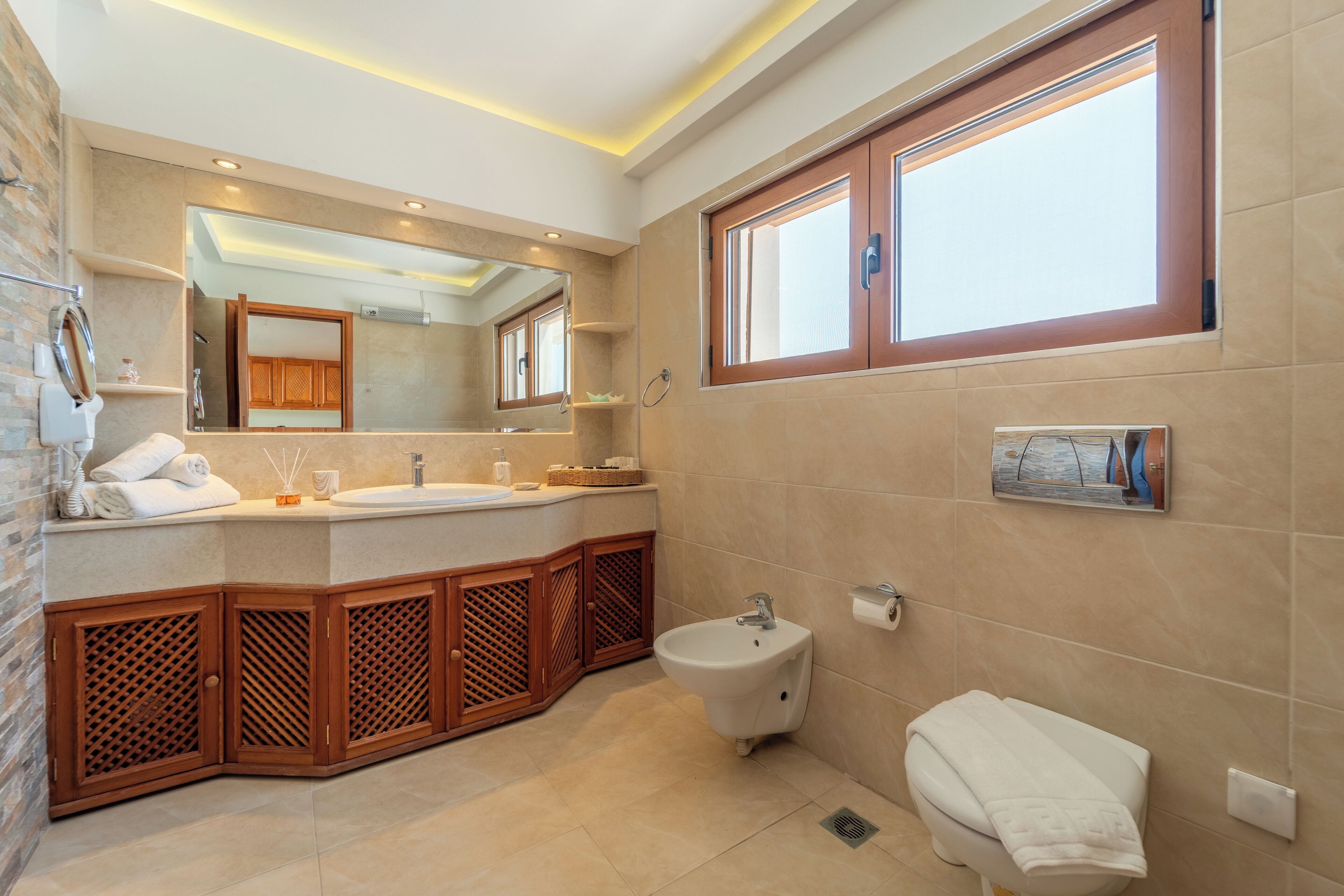 A modern bathroom with a large mirror, wooden cabinets, a bidet, and a window.