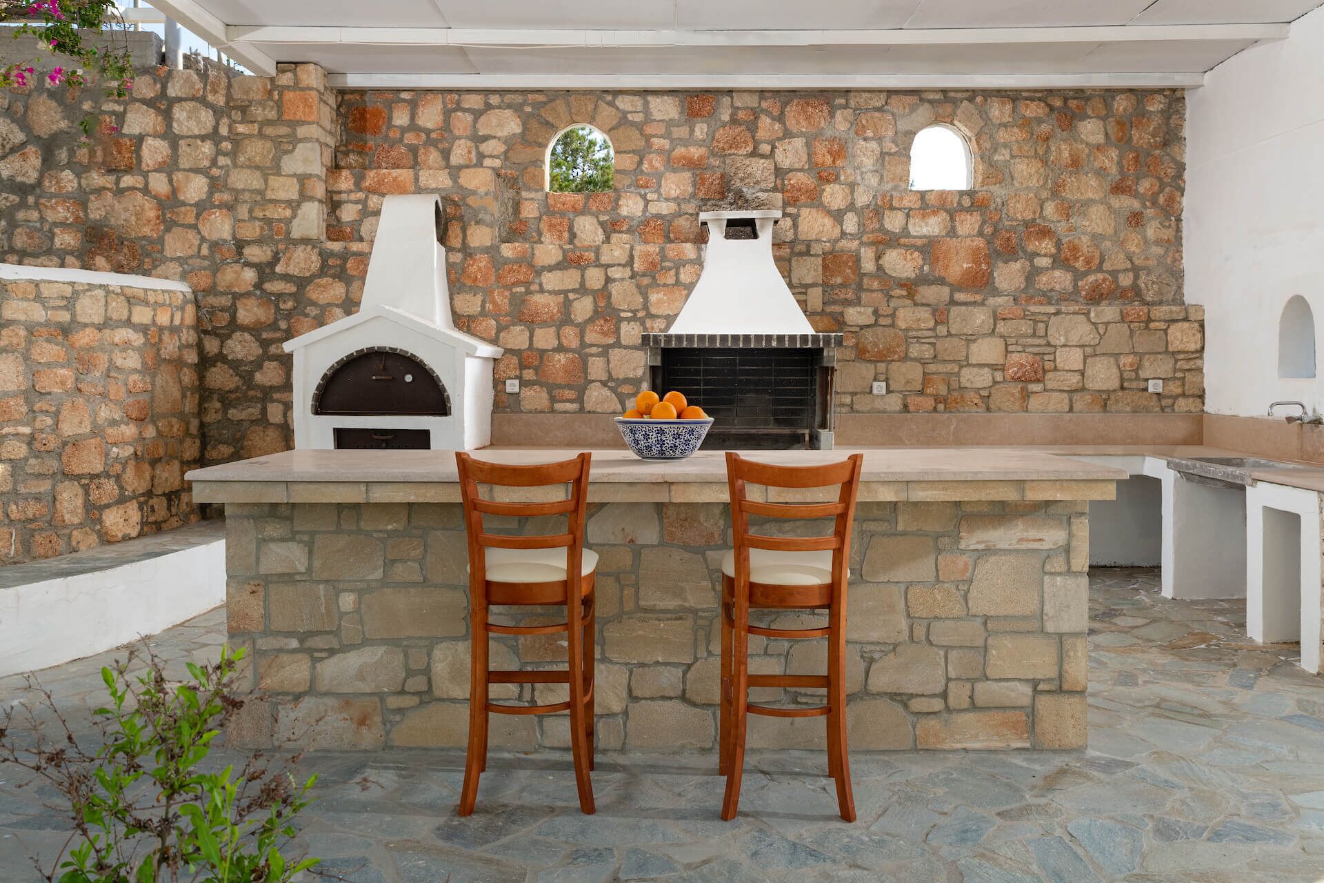An outdoor kitchen area with stone walls, two wooden bar stools, and a bowl of oranges on the counter.