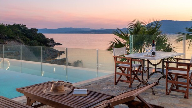 A poolside area with a table and chairs, overlooking a body of water and hills. - Arte Pefki Beachfront Suites (Rhodes)