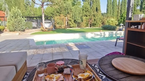 An outdoor dining area with a pool view, wooden furniture, and a table set with breakfast items.