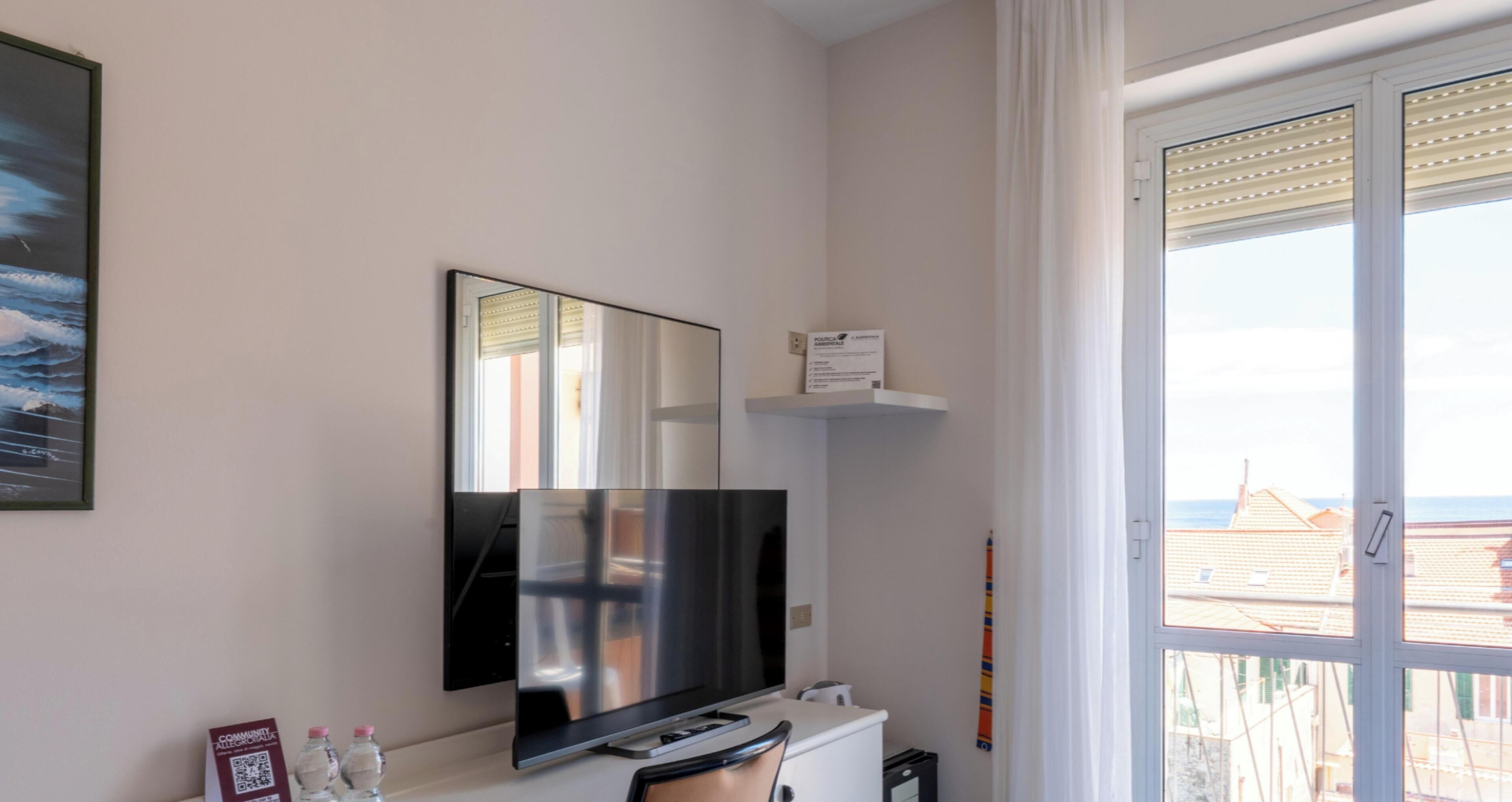Een kamer met een wandgemonteerde flatscreen televisie, een tafeltje met een afstandsbediening, en een raam met uitzicht op gebouwen en een wateroppervlak.
