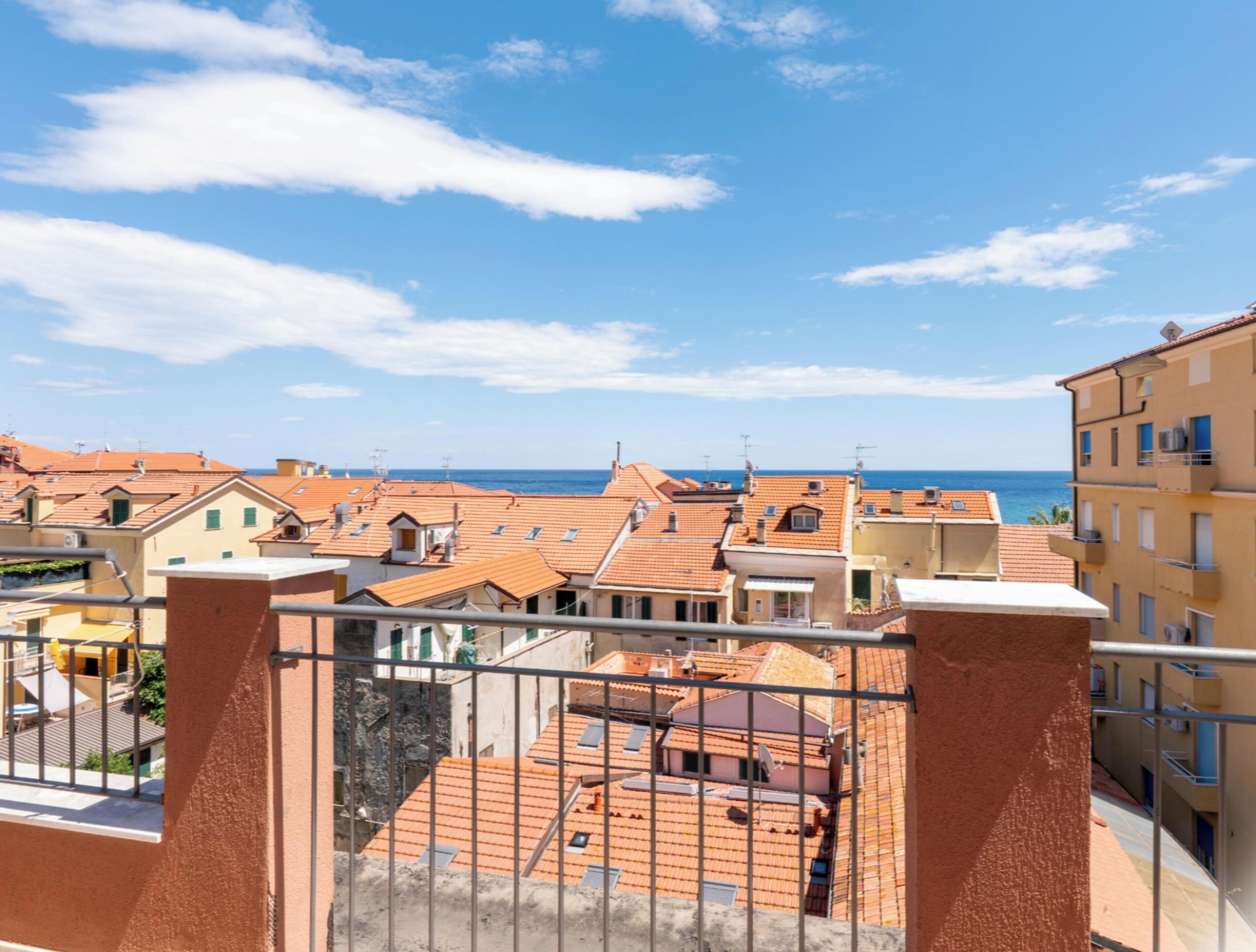 A residential area with terracotta-roofed buildings, a clear blue sky, and a view of the sea.