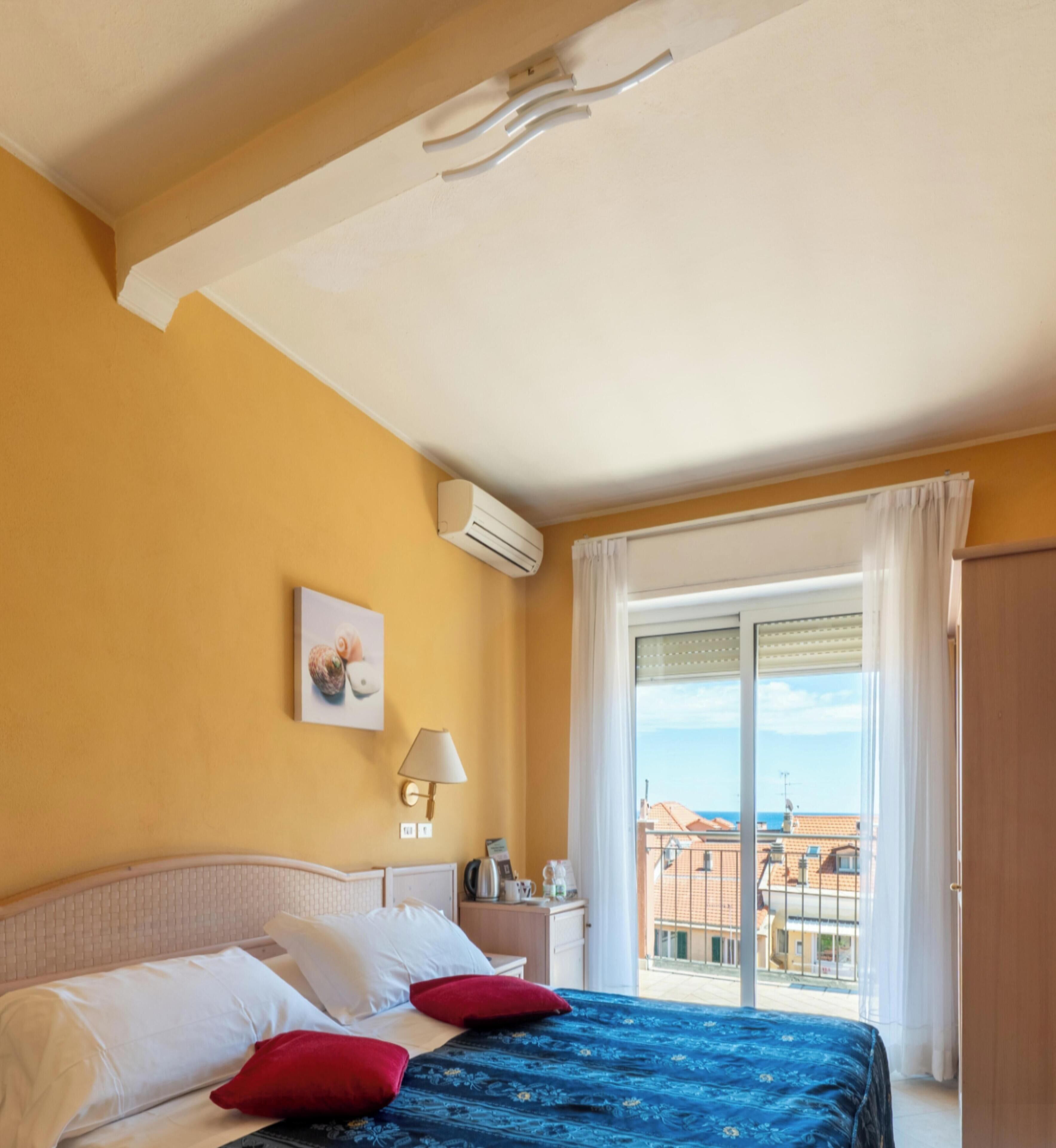 Habitación de hotel con cama, mesita de noche, lámpara, unidad de aire acondicionado y vista a edificios a través de una ventana.