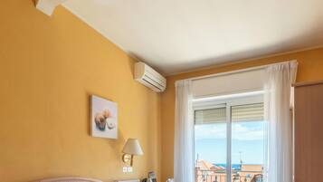 Habitación de hotel con cama, mesita de noche, lámpara, unidad de aire acondicionado y vista a edificios a través de una ventana.