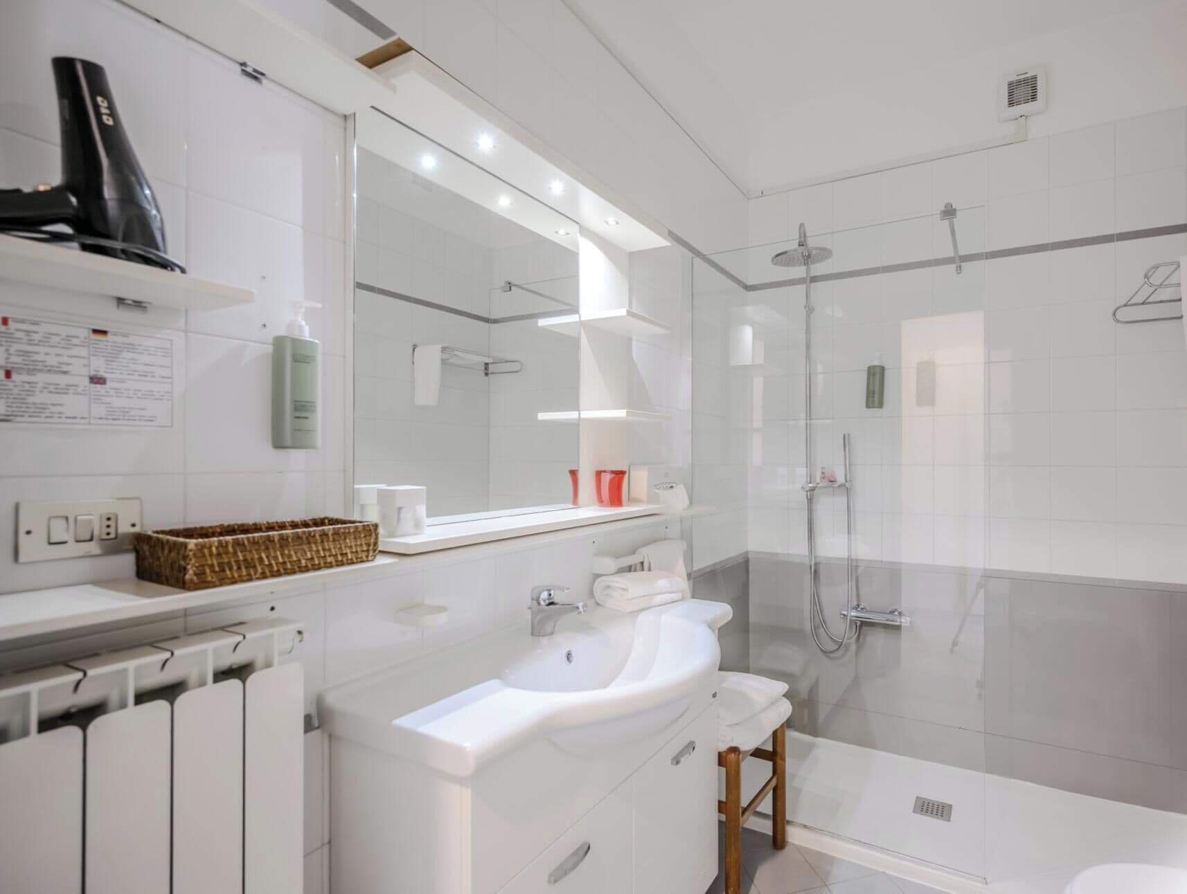 A modern bathroom with a large mirror, two sinks, a shower, and a radiator.