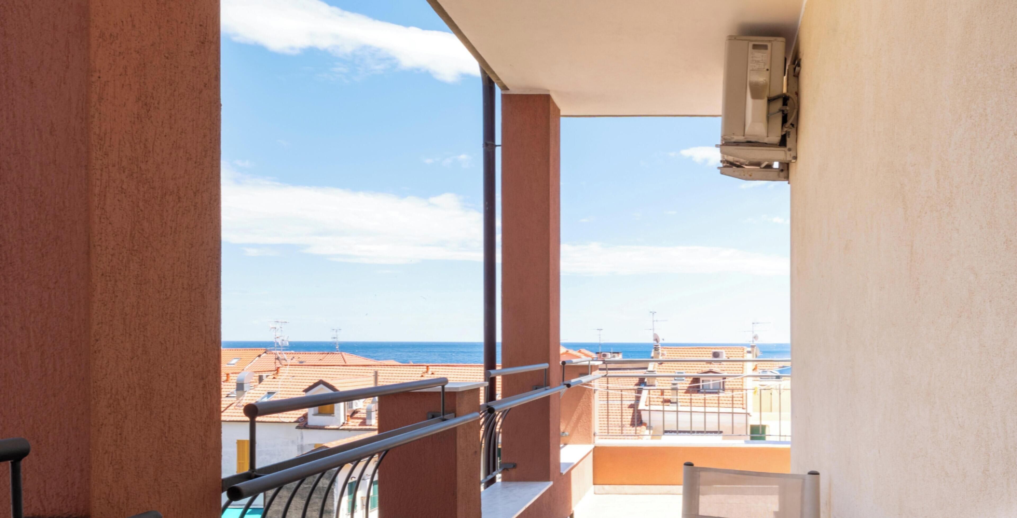 A balcony with a metal railing, overlooking a coastal area with buildings and the sea.