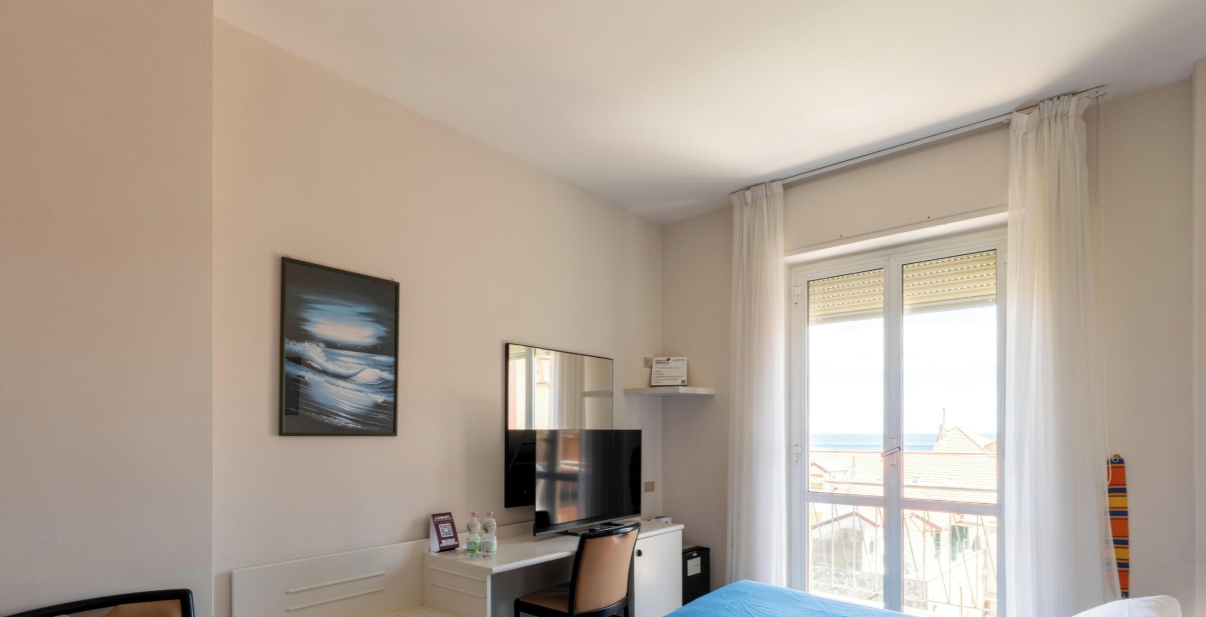 A hotel room with a bed, a desk with a TV, a chair, a window with curtains, and a painting on the wall.
