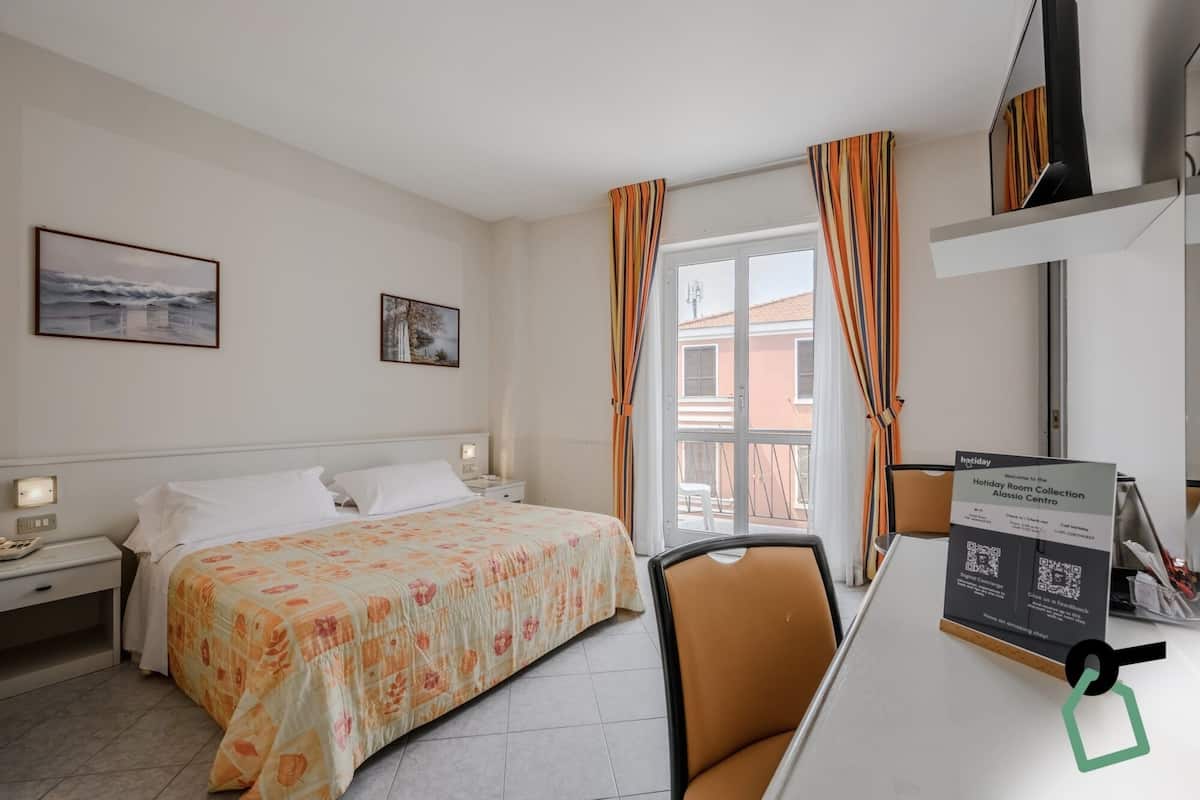 A hotel room with a bed, a desk, a chair, a window with curtains, and a sign on the desk.