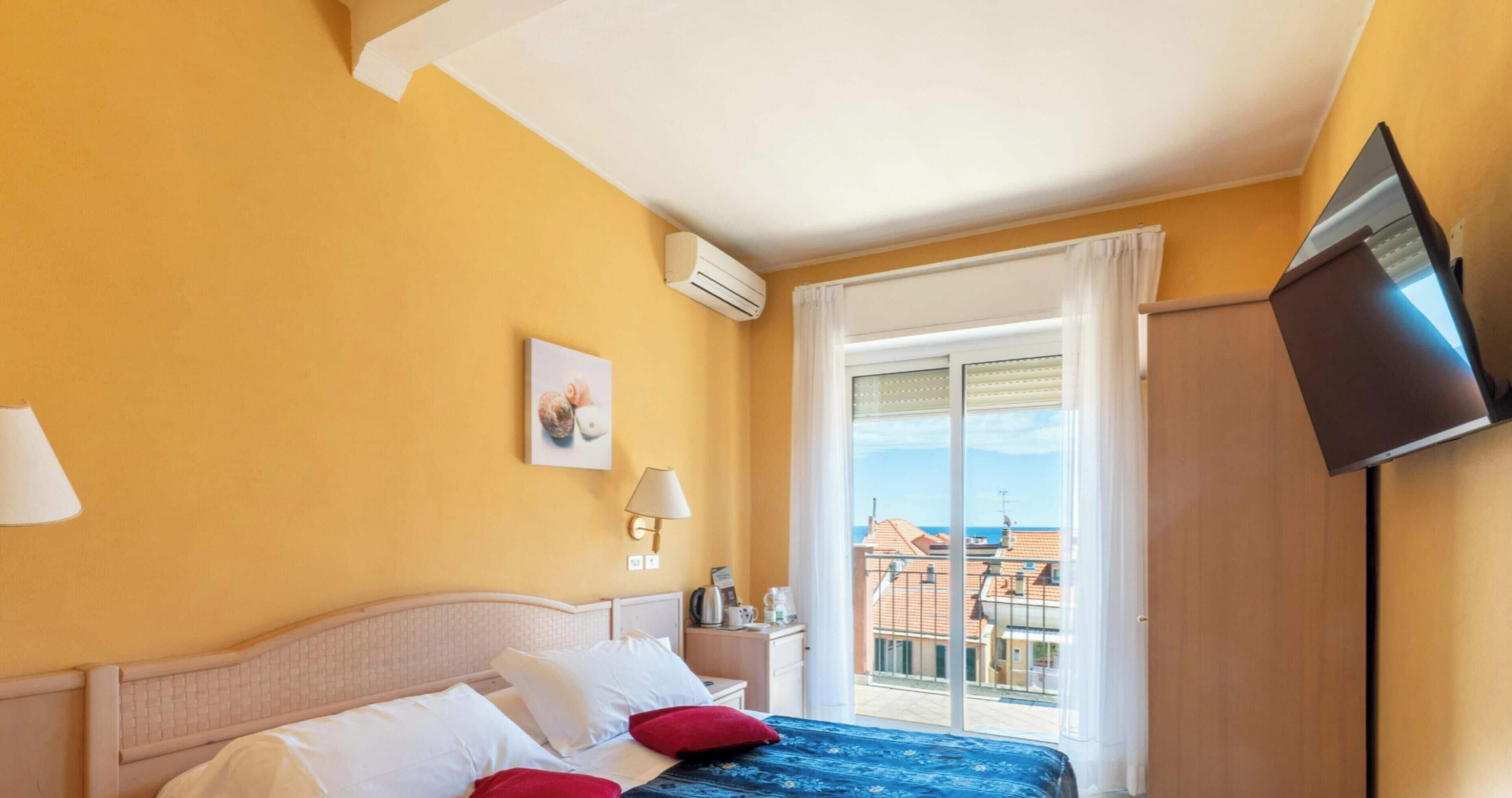 A hotel room with a bed, a TV, a window with a view of buildings, and an air conditioning unit.