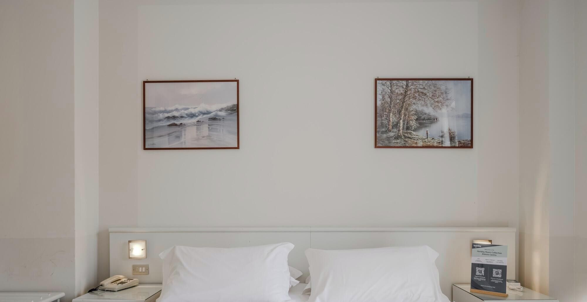 A hotel room with a bed, two framed pictures on the wall, a bedside table with a lamp, and a nightstand with a phone.