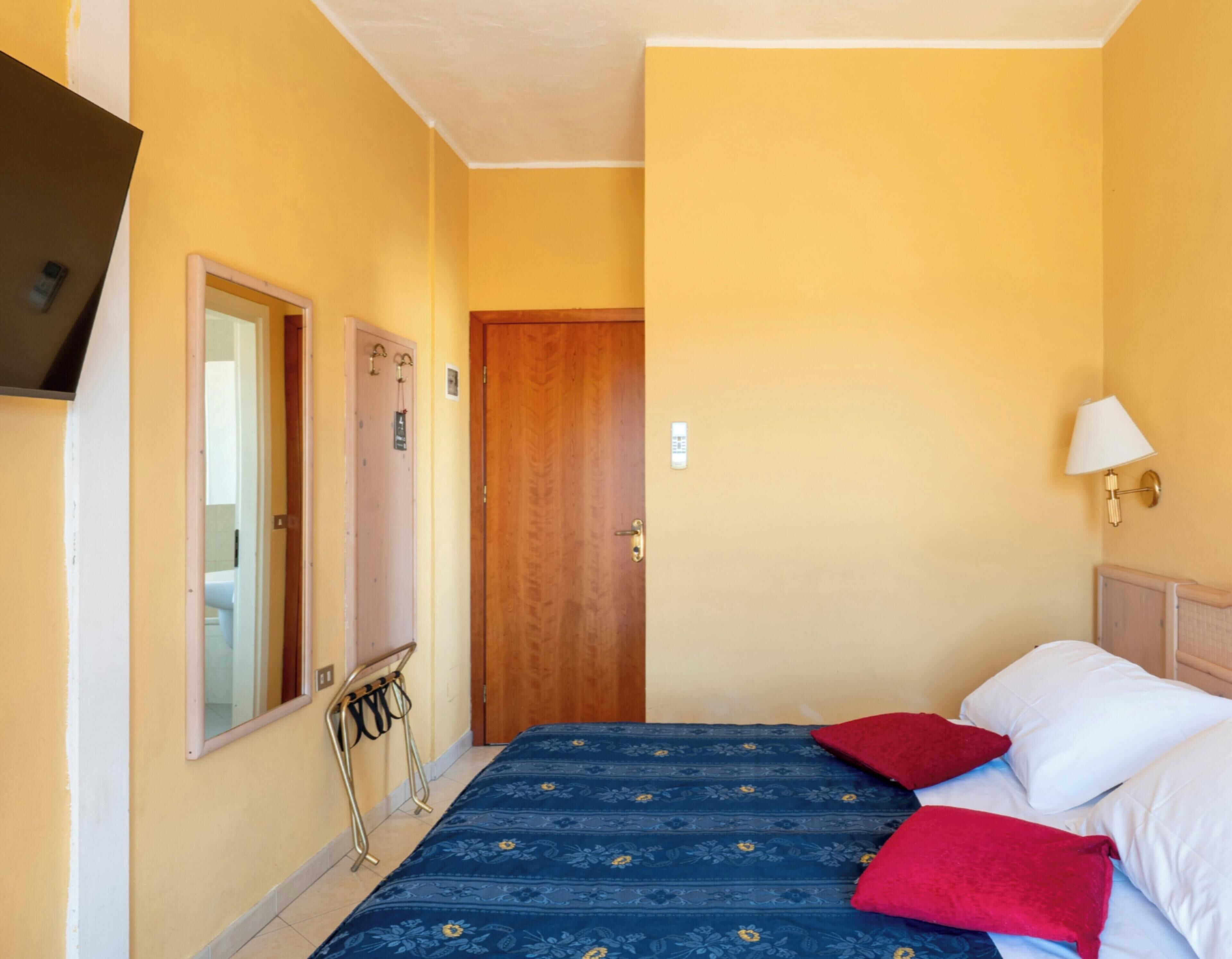 A room with a bed, a wooden door, a mirror, a wall-mounted lamp, and a small table with a chair.
