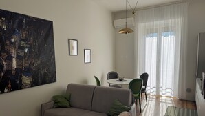 En stue med sofa, spisebord med stole, et stort maleri og et vindue med gardiner.