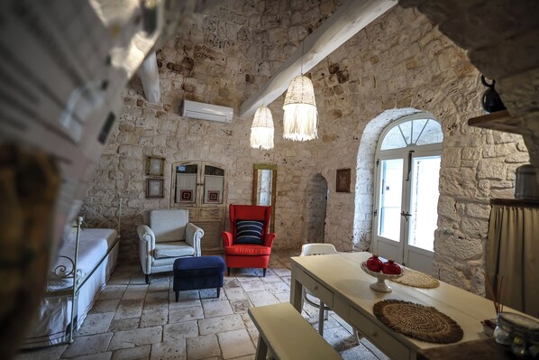 A stone-walled room with a bed, two chairs, a table with a fruit bowl, and a hanging light fixture.